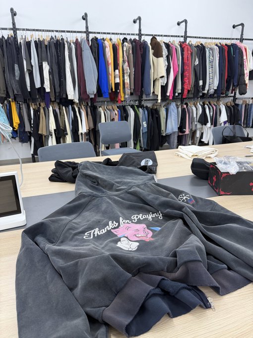 A clothing retail or warehouse setting with racks of hoodies and apparel in the background, and a dark grey hoodie on the table in the foreground featuring the embroidered text 'Thanks for Playing' with a pink character graphic.