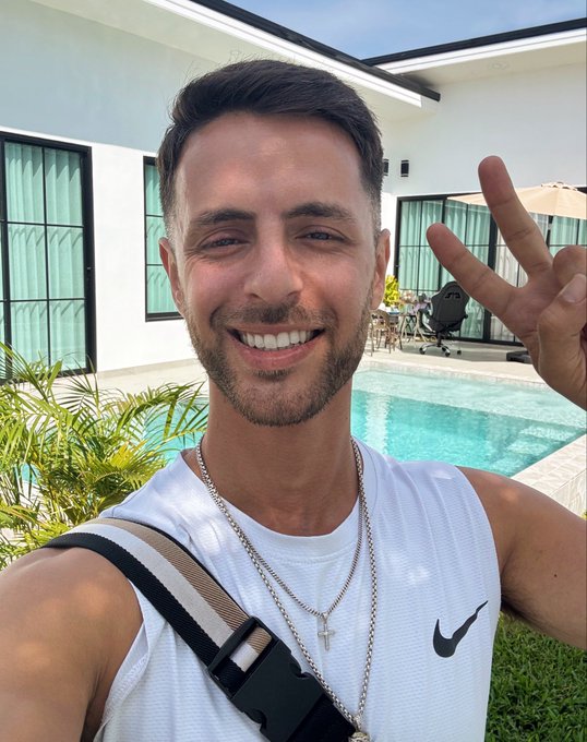 A selfie of a smiling man wearing a white Nike tank top, layered silver chain necklaces, and a crossbody bag strap, posing with a peace sign near a swimming pool in what appears to be a tropical resort setting.