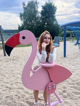 A young woman with red hair poses with a large pink flamingo inflatable chair at a sandy beach or playground area.