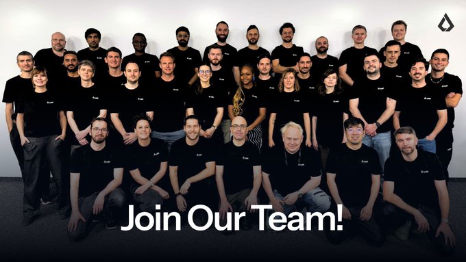 A team photo of around 30 people wearing black t-shirts posing in an office, with the text 'Join Our Team!' and a logo in the upper right corner.