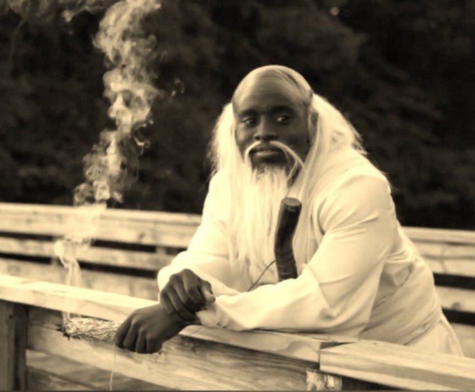 A sepia-toned portrait of a bald, bearded man in white robes smoking a long pipe, leaning against a wooden railing with smoke curling upward.