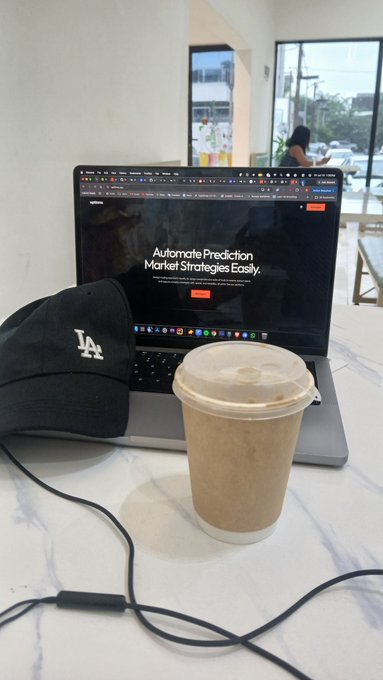 A laptop on a marble table displays "Automate Prediction Market Strategies Easily" alongside a black LA Dodgers cap, earphones, and an iced coffee.