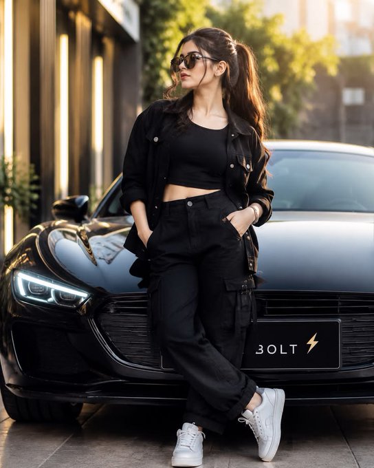 A young woman in a black crop top, jacket, and cargo pants stands in front of a black sports car, with a visible front grille badge reading '30LT' and a lightning bolt symbol.