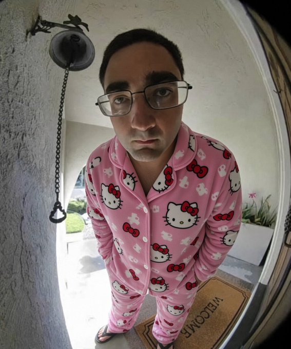 A man wearing pink Hello Kitty pajamas and glasses standing on a welcome mat at a doorway, captured through a fisheye lens.