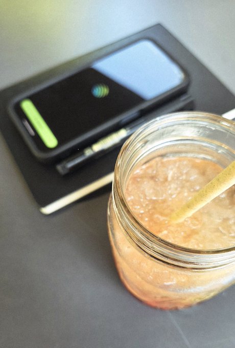A glass mason jar containing a pink/beige smoothie with a yellow straw, sitting on a gray surface next to a black notebook with a smartphone displaying a green interface on top.