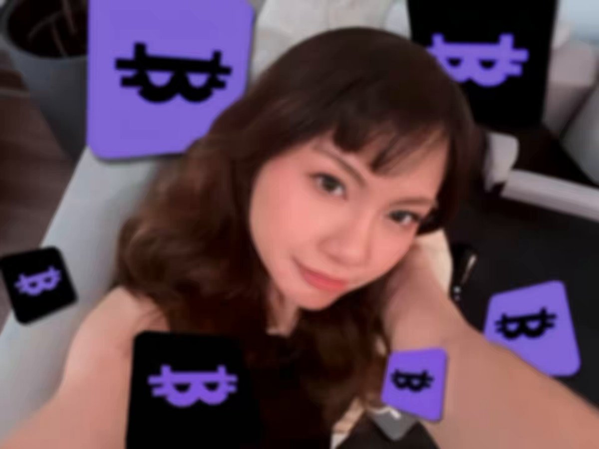 The image features a woman with brown hair wearing a black top, posing for a photo. She has her arms around herself as if she's hugging someone or something. The background of the image includes several purple squares scattered throughout, which appear to be Bitcoin logos. These logos are placed at various positions in the image, creating an interesting visual effect and adding depth to the scene.