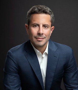A professional headshot of a man in a navy suit against a dark background, visually accompanying Saifedean Ammous's quote about inflation and Bitcoin as a solution to currency devaluation.