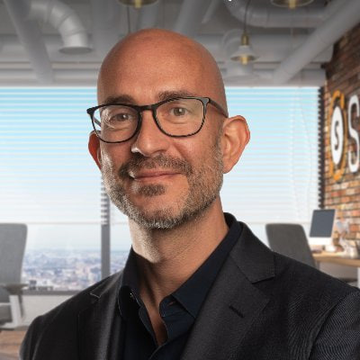 A professional headshot of a bald man with glasses, a gray beard, and a dark blazer, photographed in a modern office setting with brick walls and exposed ceiling ducts.