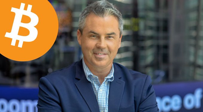 A professional headshot of a man in a navy blazer and checkered shirt with a large orange Bitcoin logo in the upper left corner.