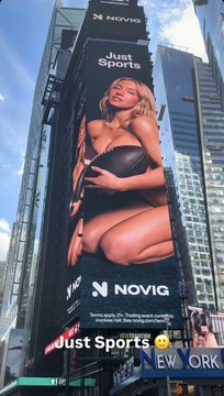 A large outdoor billboard advertisement in an urban setting for 'NOVIG' featuring a blonde woman in a black top and white shorts with the tagline 'Just Sports' and a yellow emoji sticker placed over part of the text.