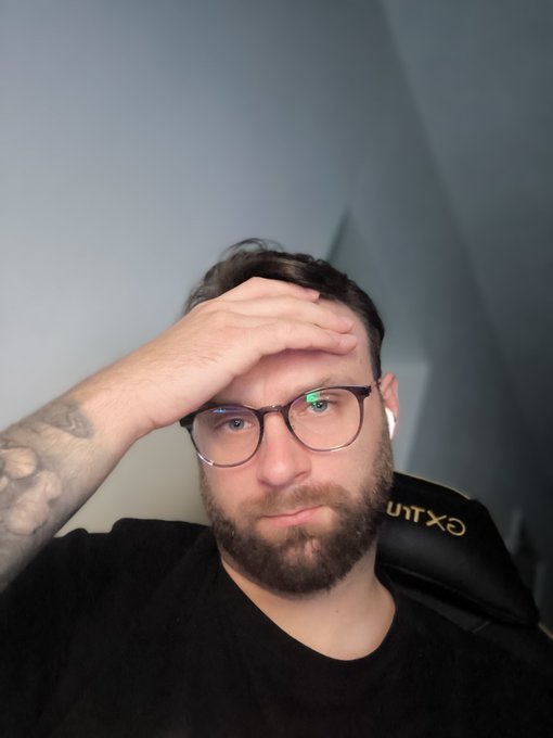 The image features a man wearing glasses and holding his head with both hands over his eyes as he looks directly at the camera. He has a tattoo visible on one of his wrists, adding a personal touch to his appearance. The background of the photo appears to be gray, giving it a neutral tone that allows the subject's facial expression and body language to take center stage in the image.