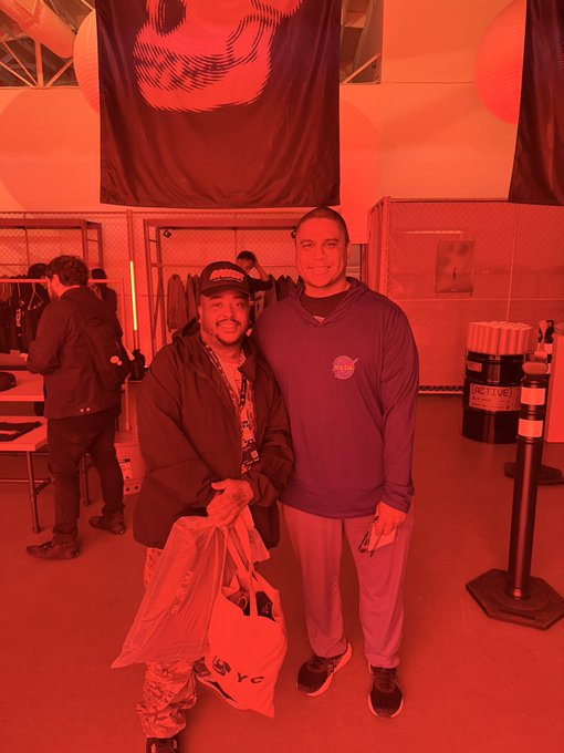 Two men pose for a photo at what appears to be an indoor event with red lighting, banners, and merchandise displays in the background.