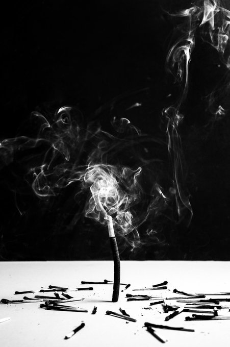 A black and white photograph of a bent incense stick emitting swirling smoke, surrounded by scattered black sticks on a white surface.