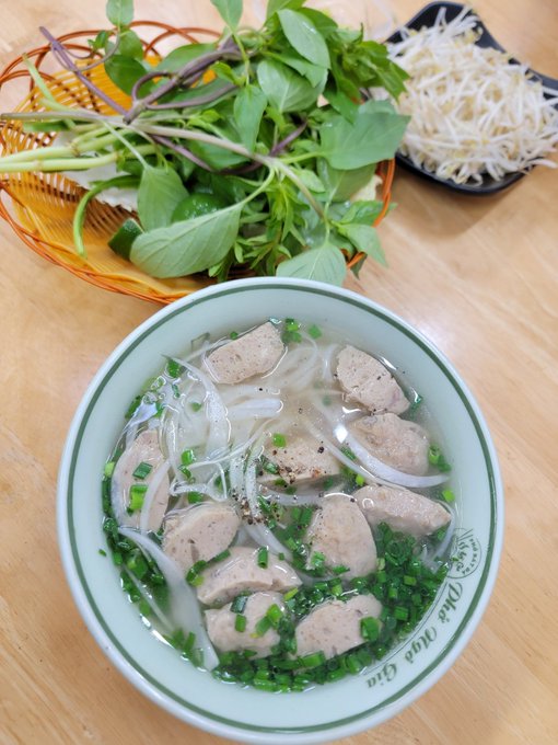 A bowl of Vietnamese soup with meatballs, rice noodles, and bean sprouts, accompanied by a basket of fresh herbs on a wooden table.