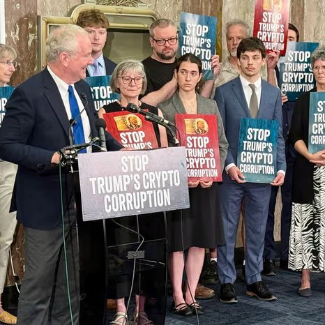 The image shows a press conference where a group of people, including a man speaking at a podium, are holding signs reading "STOP TRUMP'S CRYPTO CORRUPTION," aligning with the accompanying post about Democratic opposition to the CLARITY Act over ethics concerns regarding government officials' crypto holdings.