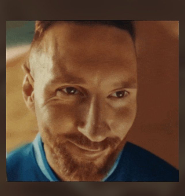 Close-up selfie photo of a smiling man with a beard wearing a blue shirt, with warm orange lighting in the background.