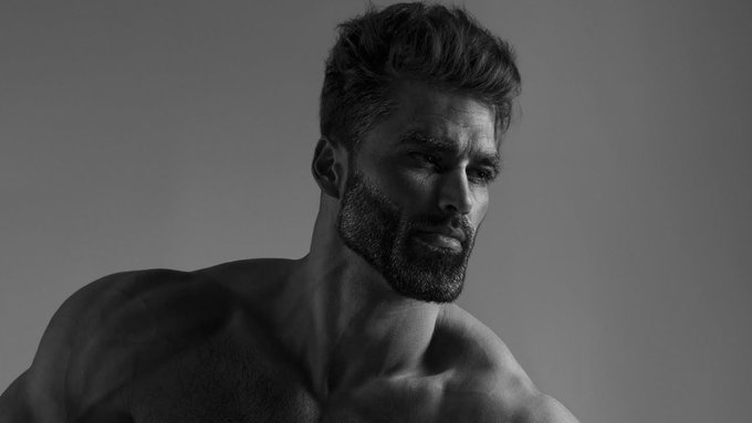 A black and white portrait of a muscular man with a beard and styled hair, looking off-camera.