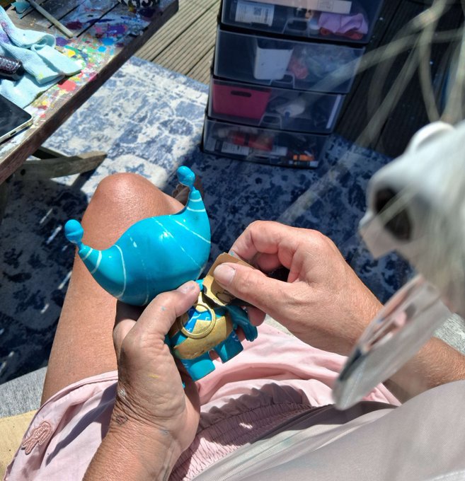 A person's hands outdoors holding and working on small blue designer toy/figurine pieces, with storage drawers and a paint-splattered table visible in the background.