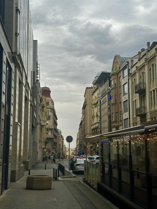 A narrow European city street lined with ornate, multi-story buildings under an overcast sky, featuring parked cars along the sidewalk and a modern glass structure on the right.