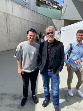 Three men stand outdoors in a casual setting, with the man in the center wearing a black blazer over a dark shirt and blue jeans.