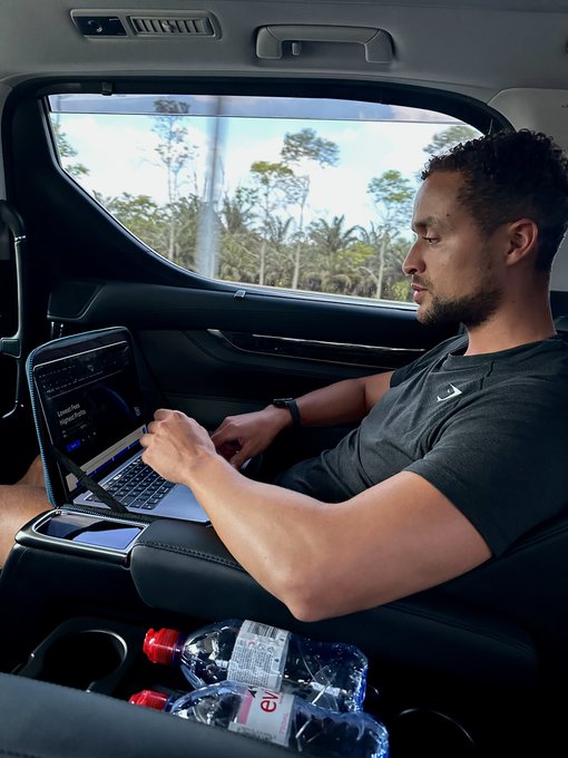 The image shows a man sitting inside a car with his laptop open in front of him. The man appears to be focused on using his laptop, possibly working or browsing the internet. There are two water bottles placed next to him, suggesting that he might be spending some time in the car and needs to stay hydrated. Additionally, there is a bottle of soda nearby, which could also indicate that the man is taking a break from work or other activities and enjoying a drink while using his laptop.