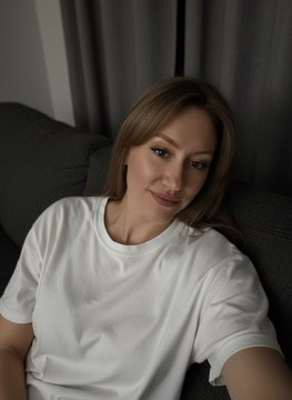 A young woman with long brown hair, wearing a white t-shirt, takes a casual selfie while sitting on a dark couch, smiling softly at the camera. The relaxed, intimate vibe of the photo complements her morning greeting, promoting the release of her new 1/1 NFT collection on @objktcom.