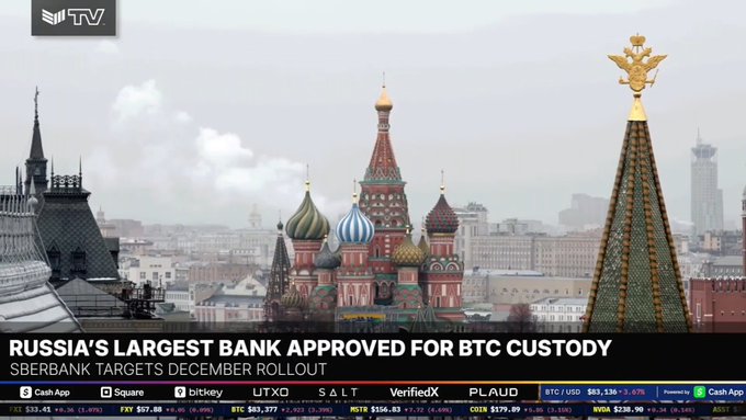 A news broadcast shows St. Basil's Cathedral in Moscow with the headline 'RUSSIA'S LARGEST BANK APPROVED FOR BTC CUSTODY' and 'SBERBANK TARGETS DECEMBER ROLLOUT,' alongside cryptocurrency tickers.