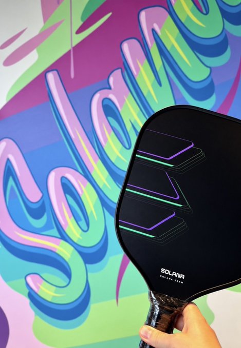 Photo of a Solana-branded pickleball paddle being held in front of a colorful 'Solana' graffiti mural.
