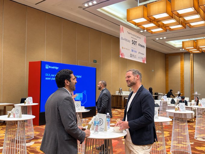 Two men in business attire conversing and smiling at a corporate event, with a 'Thank You to Our Sponsors GLADSCALE SOT GRANITE' banner visible and a Broadridge digital display screen in the background.