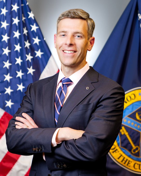 A smiling man in a dark suit with a blue and orange striped tie stands with arms crossed in front of an American flag and what appears to be an Office of the Comptroller of the Currency (OCC) seal, suggesting his role in the agency's recent push to revitalize new bank chartering for digital assets and emerging technology firms.