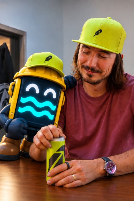 A man wearing a yellow cap smiles while opening an energy drink next to a small yellow robot toy with a digital screen face displaying a wavy pattern.