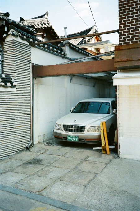 A silver sedan parked in a narrow driveway between a traditional Korean tiled-roof building and a brick wall, with a green Korean license plate.