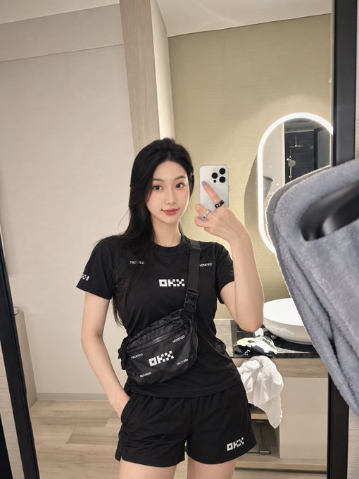 A young woman takes a mirror selfie in a hotel bathroom, wearing an all-black outfit (t-shirt, shorts, crossbody bag) branded with the 'OKX' cryptocurrency exchange logo, while holding a smartphone.