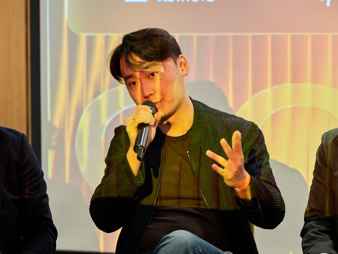 A man in a dark jacket speaks into a microphone while gesturing with his hand at what appears to be a panel discussion or conference, seated between two other individuals. The setting aligns with the post's context of a financial discussion featuring Haonan Li of @CodexFX addressing the GENIUS Act's role in legitimizing stablecoins.