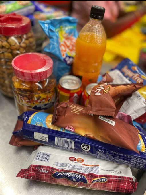 An assortment of snacks and grocery items on a table, including a bottle of orange Fanta, various chip bags (Valentine's and others), a jar of nuts, canned goods, and packaged foods.