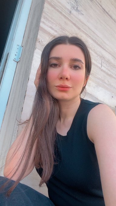 The image features a woman with long brown hair wearing a black tank top, sitting against a white wall or door frame. The photo has been edited by someone named Gm Artist, who has added a caption that reads "Gm Artist/ Art (“Held by Yesterday” 1/1_ 0.11 $ETH)". This caption suggests that the image was taken and edited by an artist for a website or social media platform.