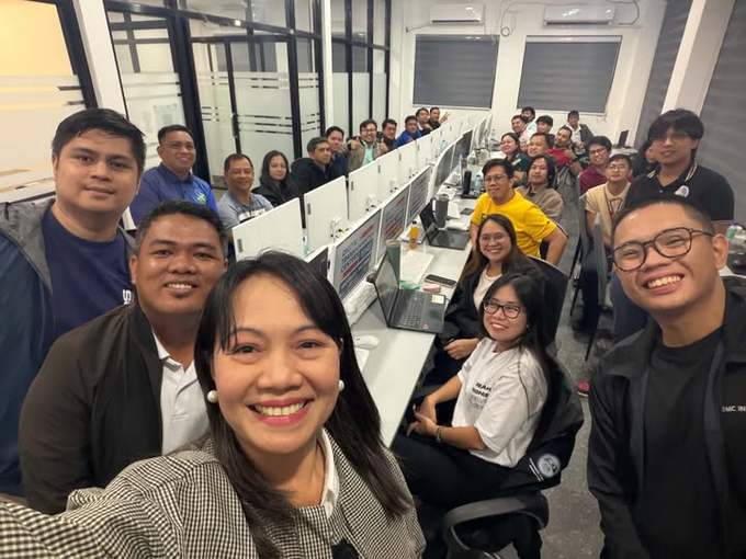 A group selfie of smiling team members in an office, with a woman in the foreground and colleagues standing and seated at desks with monitors in the background.