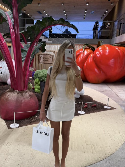 A blonde woman in a white dress takes a mirror selfie with a phone, holding a "MONSTER" shopping bag, in front of a colorful farm stand display featuring a giant pumpkin and beet. A sign reading "FARM FRESH" is visible in the background, complementing the post's playful "what is KBW" theme by evoking a wholesome, rustic aesthetic.