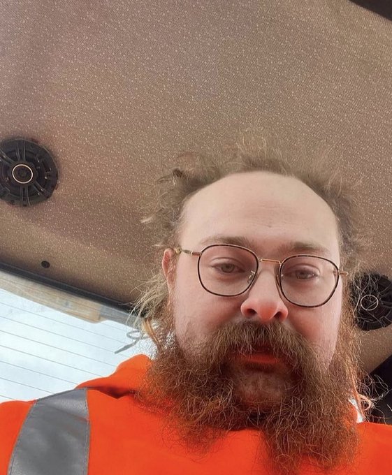 A low-angle selfie of a bearded man wearing glasses and an orange high-visibility work shirt, taken inside a vehicle with a textured ceiling visible behind him.