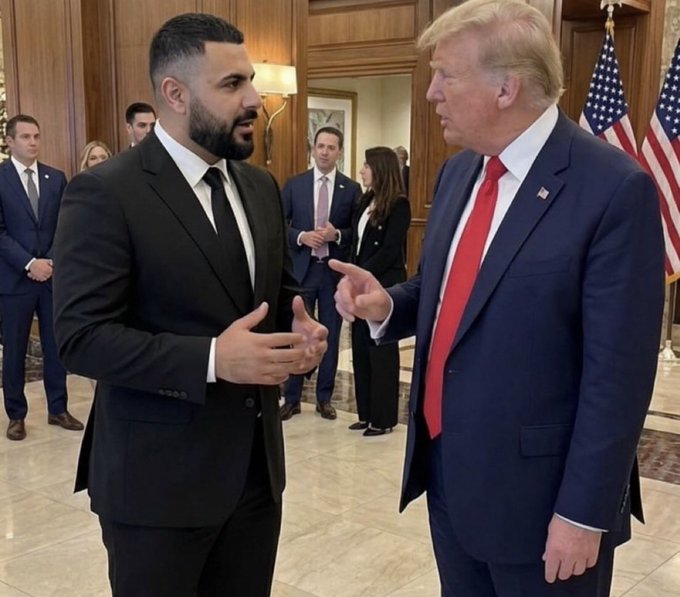 Donald Trump in a dark suit and red tie stands indoors talking with a bearded man in a black suit, with an American flag and other people visible in the background.