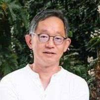 A headshot of a middle-aged man with short graying hair and glasses, wearing a white shirt against a dark green foliage background.