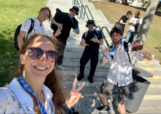A group of young people wearing matching lanyards pose for a selfie on outdoor concrete stairs, with the person in the foreground smiling and flashing a peace sign.