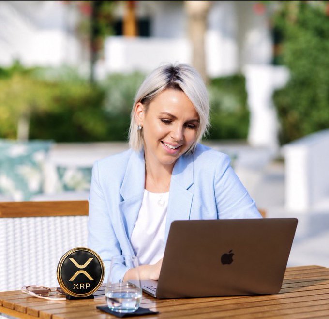 A blonde woman in a light blue blazer smiles while working on a MacBook at an outdoor table, with an XRP cryptocurrency logo coin beside her laptop.