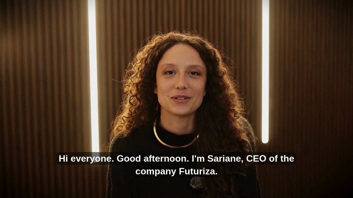 The image features a woman with curly hair wearing a black top, smiling at the camera. The background consists of brown walls that match her outfit. In the bottom right corner of the image, there are two lines of text written in white font. One line reads "Hi everyone, Good afternoon, I'm Sariane, CEO of the company Futuriza." and another line states "Together with the Embassy of Brazil and Input Output Group, we connected traditional fintech and retail companies from Brazil and Argentina, while also showcasing potential blockchain implementations for their businesses."