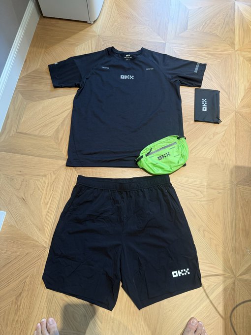 An overhead shot of a black athletic outfit consisting of a t-shirt and shorts with 'DKXX' branding, laid out on a wooden floor alongside a green waist pack and a folded item, with bare feet visible at the bottom of the frame.