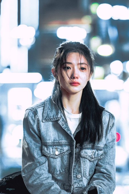 A young woman with long dark hair wearing a light blue acid-wash denim jacket and white shirt sits against a backdrop of blurred city lights at night.