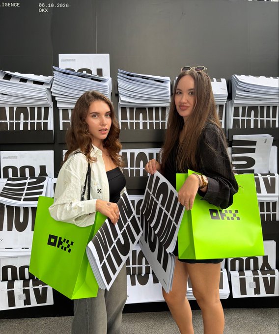 Two women pose in front of a wall displaying "OKX" branded posters, holding matching neon green OKX shopping bags and oversized black "OKX" signs. The setup reflects the OKX event mentioned in the post, where the user met Valerie (@Valerie_Onchain) while attending the branded gathering.