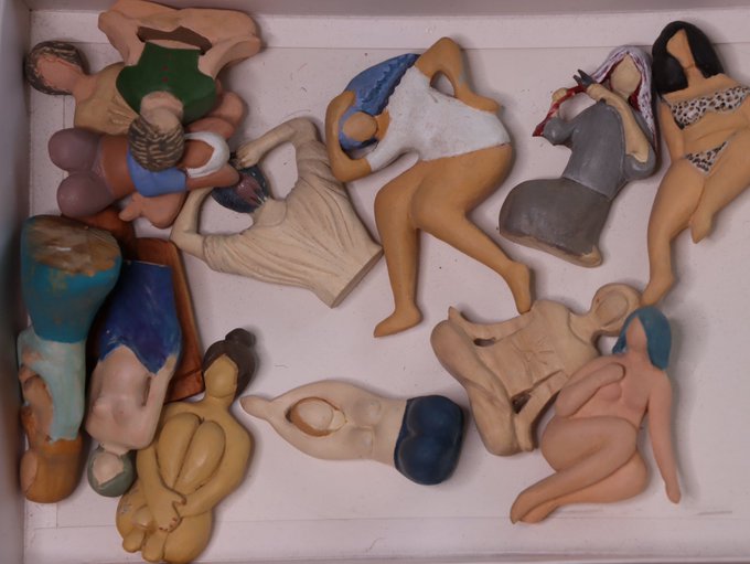 Artistic display of handcrafted ceramic figurines depicting various human figures in different poses and styles, arranged on a white shelf.
