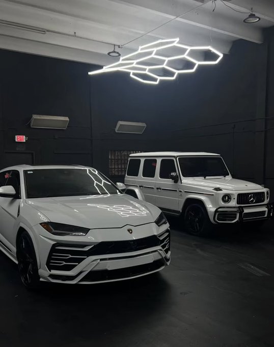 Two white SUVs—a Lamborghini Urus and a Mercedes-Benz G-Wagon—are displayed side by side in a dimly lit showroom featuring a distinctive hexagonal LED ceiling installation, perfectly capturing the "Gn bros" sentiment of elite automotive camaraderie.