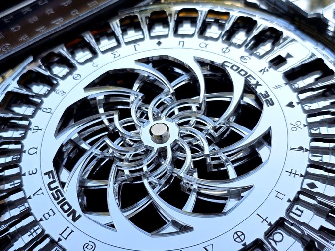 A close-up macro shot of a chrome metallic emblem featuring an intricate spiral design, with the words 'CODEX 2R' and 'FUSION' engraved around the edge alongside various small symbols.
